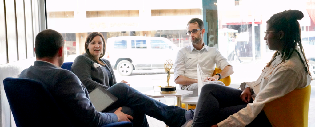Four people sit in a circle of modern lounge chairs near large windows in a bright meeting space. A small table sits between them. A street with parked vehicles can be seen through the windows.